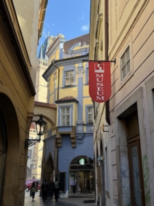 At upper right, a bright red sign promoting Prague’s Sex Museum. Dan Brown’s character known as The Golem is described as walking briskly past this point while en route to a home tucked in among the narrow streets found in the area around the Old Town Square.