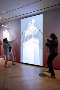 An interactive feature in “The Occupied City” exhibit at the Museum of the City of New York. On July 9, 1776, soldiers and sailors toppled a statue of King George III, located in Bowling Green. That act has inspired numerous traditional artworks, but in this case, visitors are provided physical ropes in order to “topple” a digital statue. Photo by Filip Wolak.